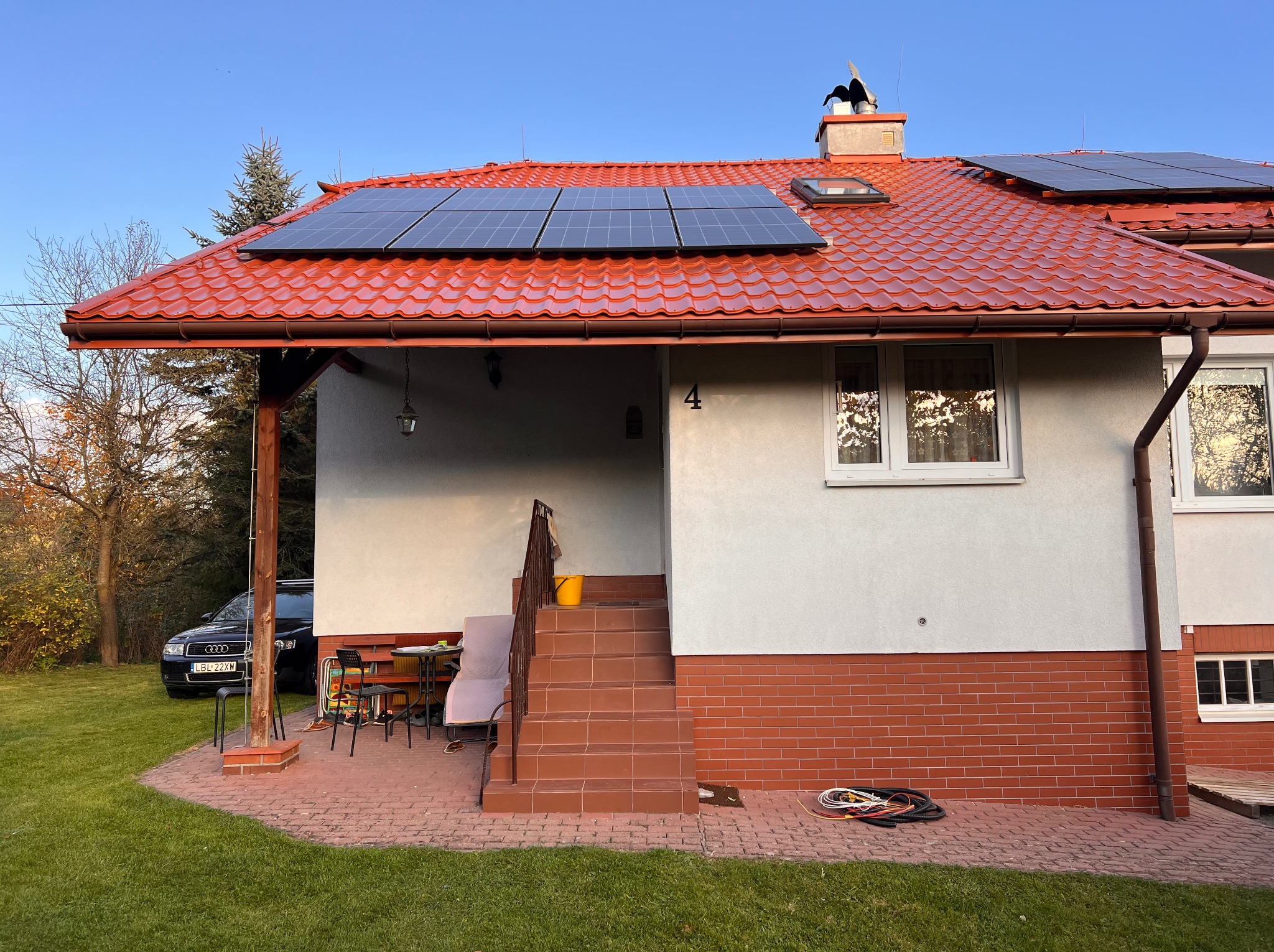 Dom jednorodzinny z czerwoną dachówką i panelami fotowoltaicznymi na dachu, widoczne schody wejściowe i częściowo ogród z samochodem w tle.