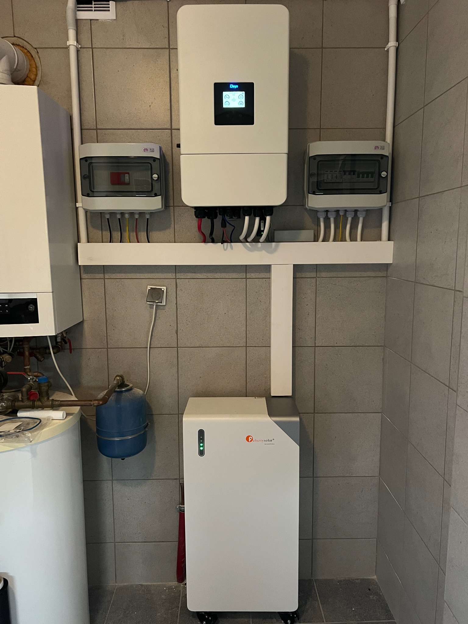 Zestaw urządzeń: inwerter, rozdzielnice i akumulator energii Felicitysolar, zamontowane na ścianie z szarych płytek. Widoczne przewody i rury instalacyjne.