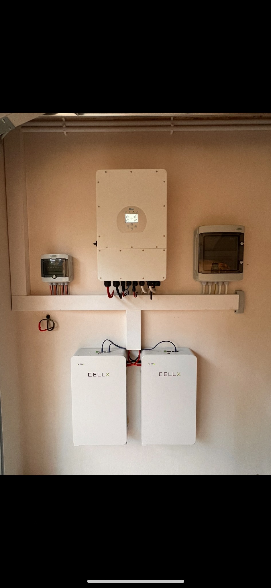 Biały inwerter i dwa akumulatory CELLX zamontowane na ścianie wewnątrz pomieszczenia. Widoczne przewody i skrzynki elektryczne. Instalacja fotowoltaiczna w domu.