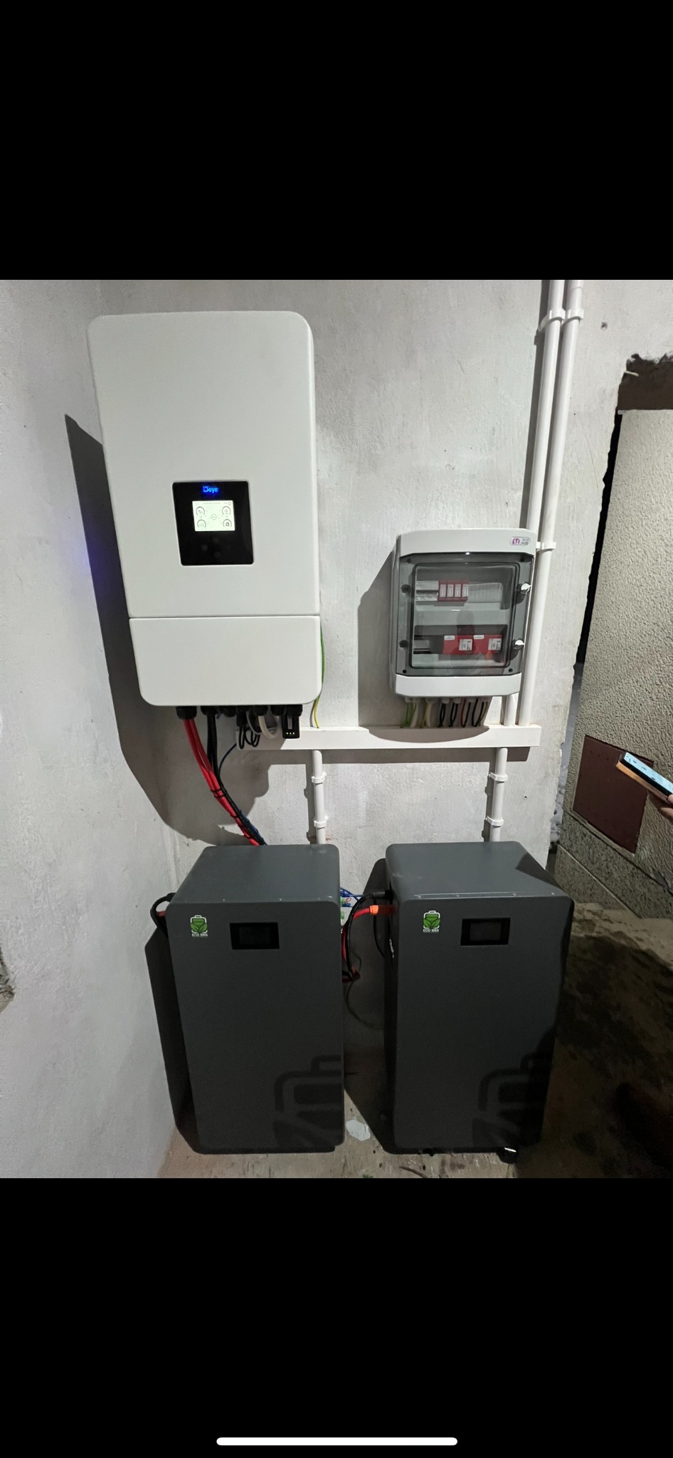 Zamontowany inwerter z dwoma akumulatorami i rozdzielnicą na ścianie. Widoczne kable i rury instalacyjne. Szara kolorystyka akumulatorów.