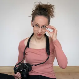 Kobieta w okularach z aparatem fotograficznym na szyi, siedząca na podłodze i poprawiająca okulary