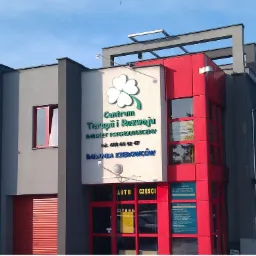 Fasada budynku Centrum Terapii i Rozwoju z logo czterolistnej koniczyny, napisem 'Gabinet Psychologiczny' i numerem telefonu, część budynku w kolorze czerwonym, część w szarym, z widocznymi...