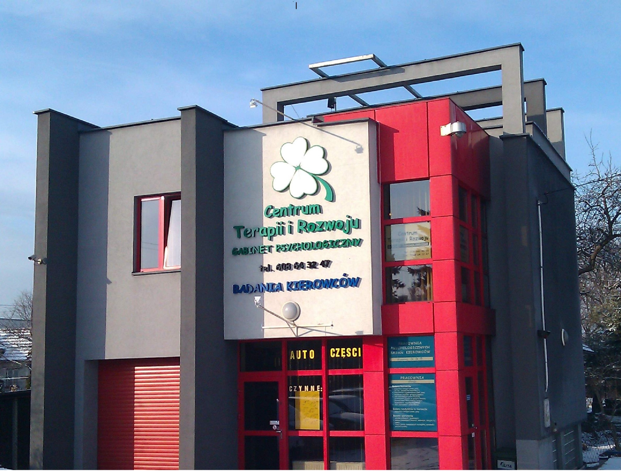 Fasada budynku Centrum Terapii i Rozwoju z logo czterolistnej koniczyny, napisem 'Gabinet Psychologiczny' i numerem telefonu, część budynku w kolorze czerwonym, część w szarym, z widocznymi...