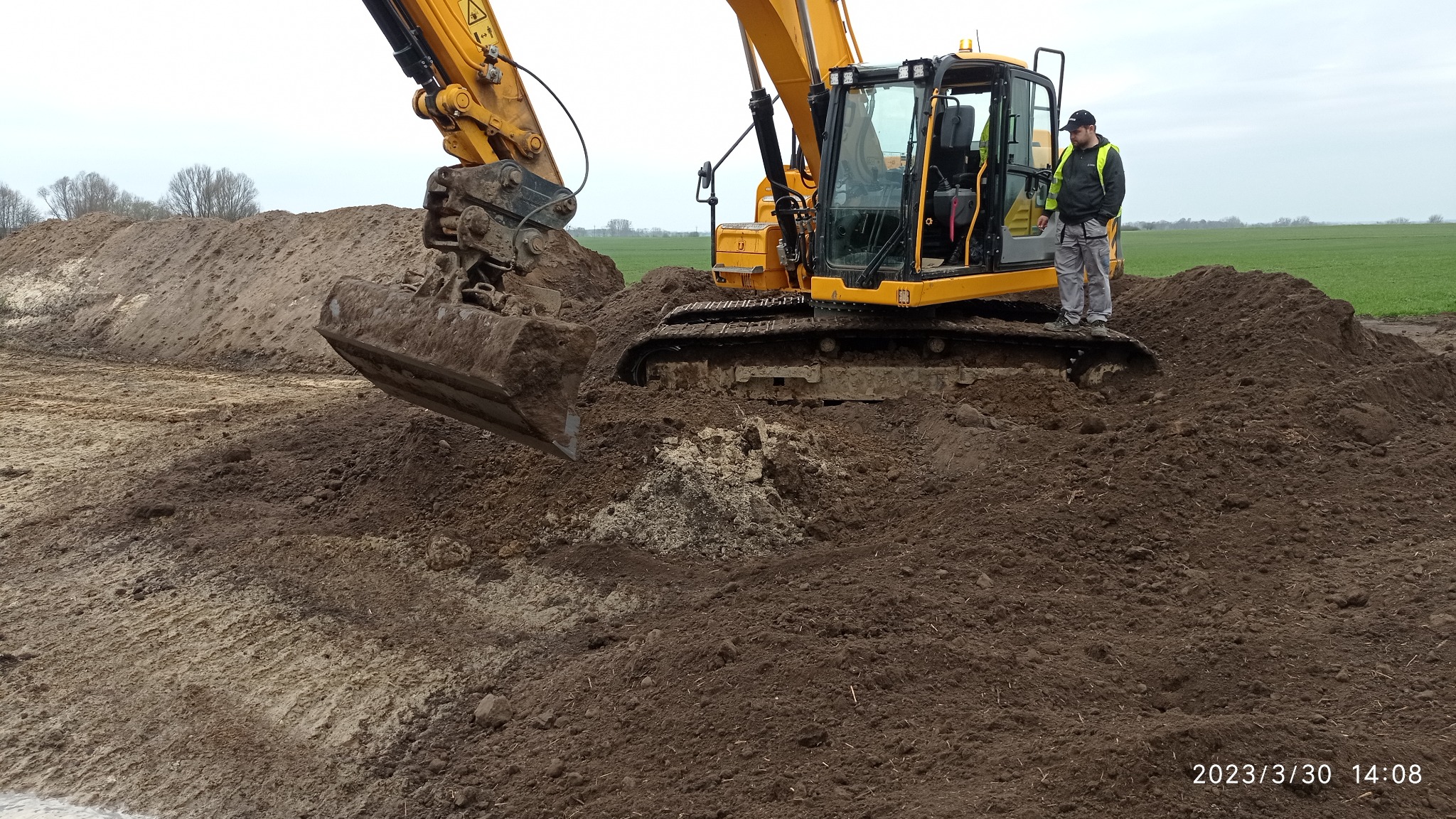 Żółta koparka gąsienicowa z łyżką, na tle pola i nieba, z operatorem w kamizelce odblaskowej stojącym na gąsienicy, pracująca na placu budowy z usypaną pryzmą ziemi.