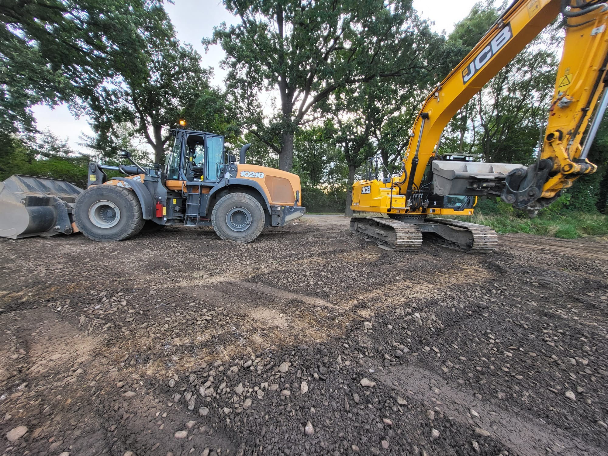Żółta koparka gąsienicowa JCB i ładowarka kołowa CASE 1021G, stojące na wyrównanym, ciemnym gruncie z widocznymi śladami opon, w tle drzewa.
