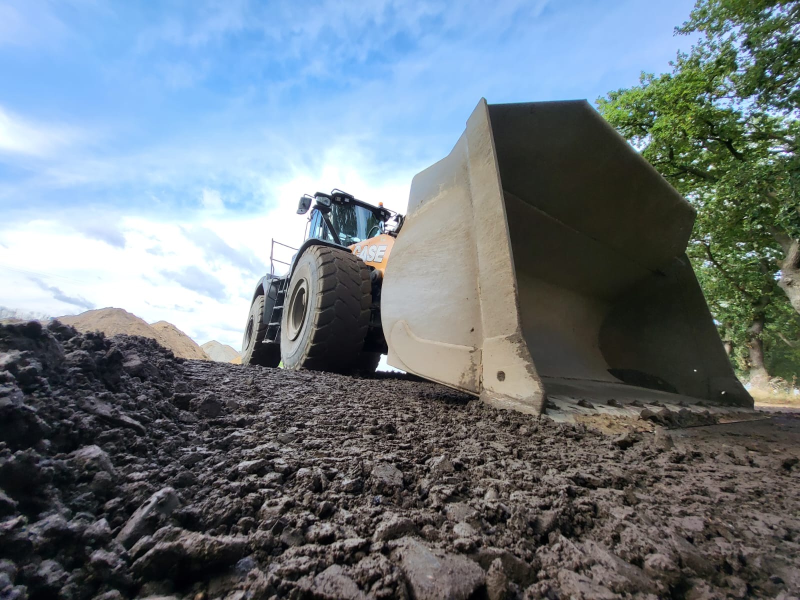Szerokokątne ujęcie ładowarki kołowej CASE z dużą łyżką na nierównym, błotnistym terenie pod błękitnym niebem z promieniami słońca.