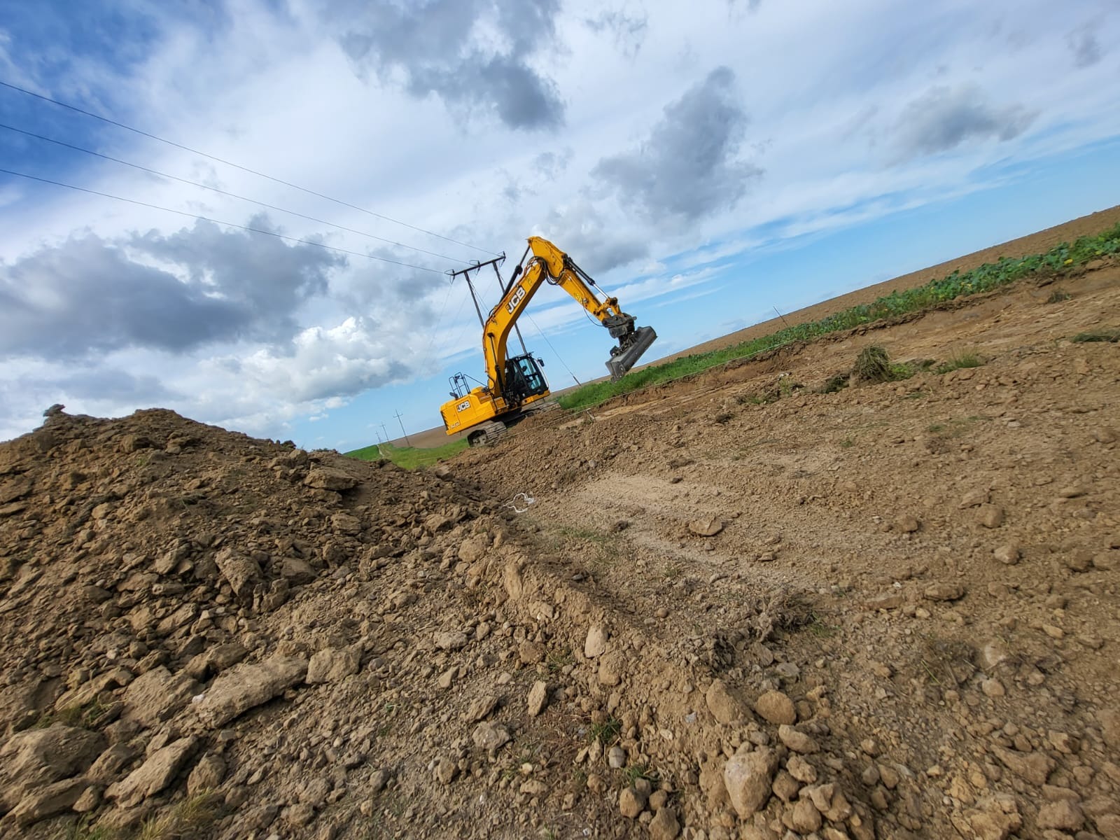 Żółta koparka JCB na nierównym terenie, przygotowuje wykop pod instalację elektryczną, widoczne słupy energetyczne i linia ziemi z roślinnością pod pochmurnym niebem.