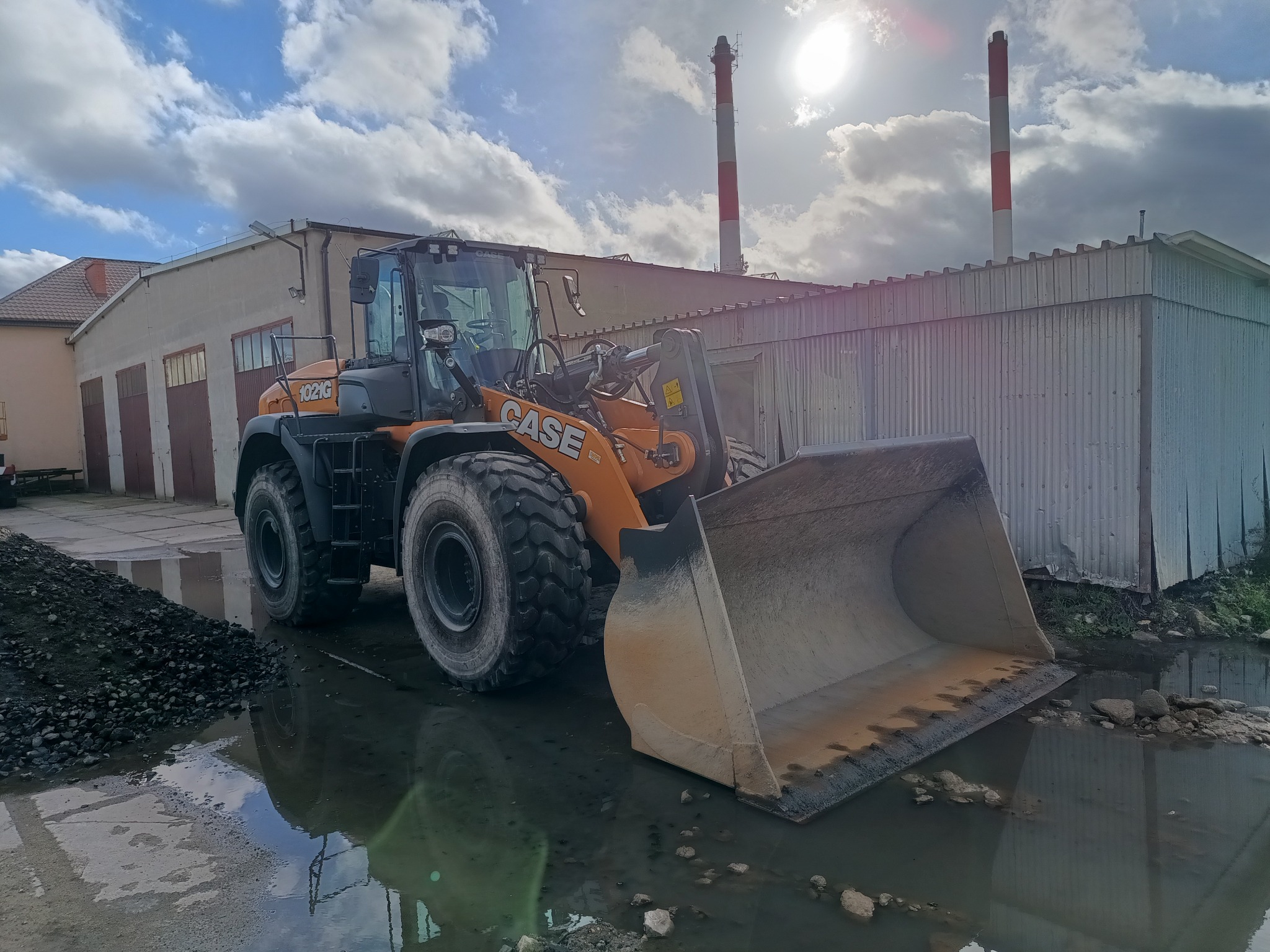 Żółto-czarna ładowarka kołowa CASE 1021G stoi na terenie budowy, z odbiciem w kałuży; w tle budynek z garażami i kominy, a nad nimi pochmurne niebo z przebijającym słońcem.
