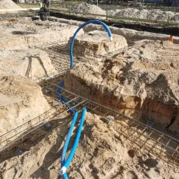 Fundamenty zbrojone stalą i niebieskie rury w wykopie w Nowych Kościeliskach, widoczni pracownicy w tle.