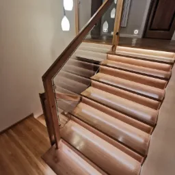 Schody drewniane z podświetleniem LED na stopniach i szklaną balustradą, widok z g&oacute;ry, z trzema wiszącymi lampami i oświetleniem podłogowym w tle.