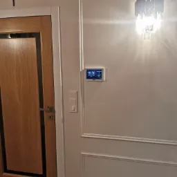 Panel sterowania alarmem domowym na ścianie z dekoracyjnymi listwami, obok drzwi z krzyżem na ścianie.