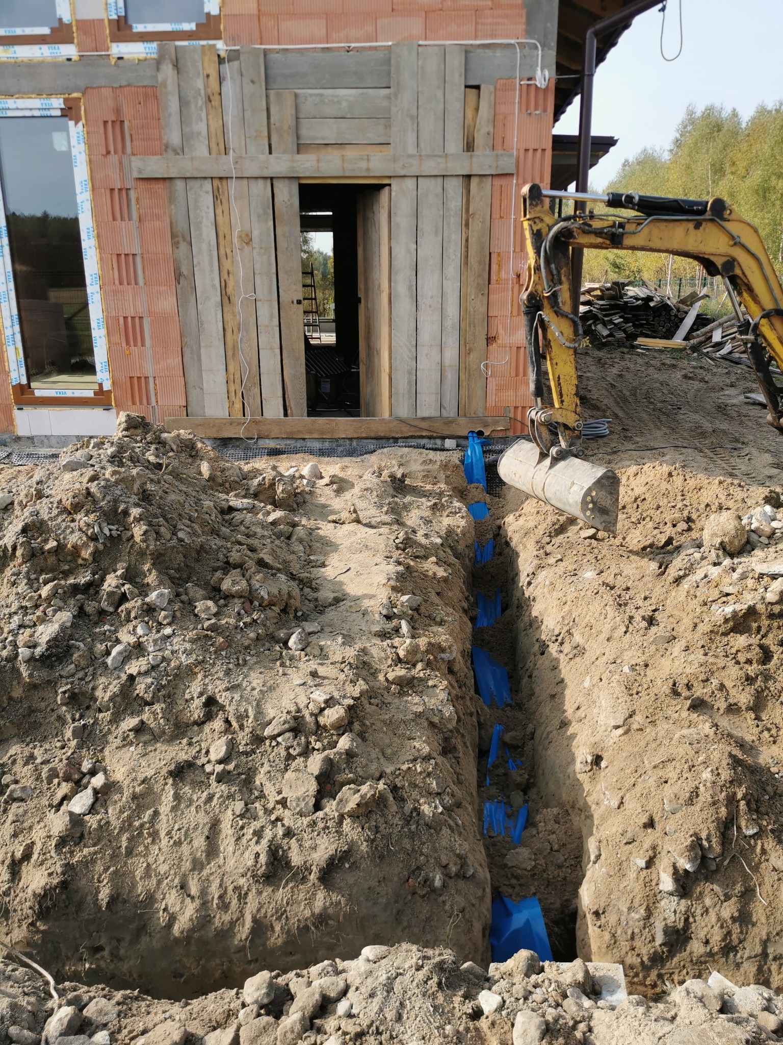 Żółta koparka wykonuje wykop pod fundament budynku z cegły, w wykopie widoczne niebieskie rury.
