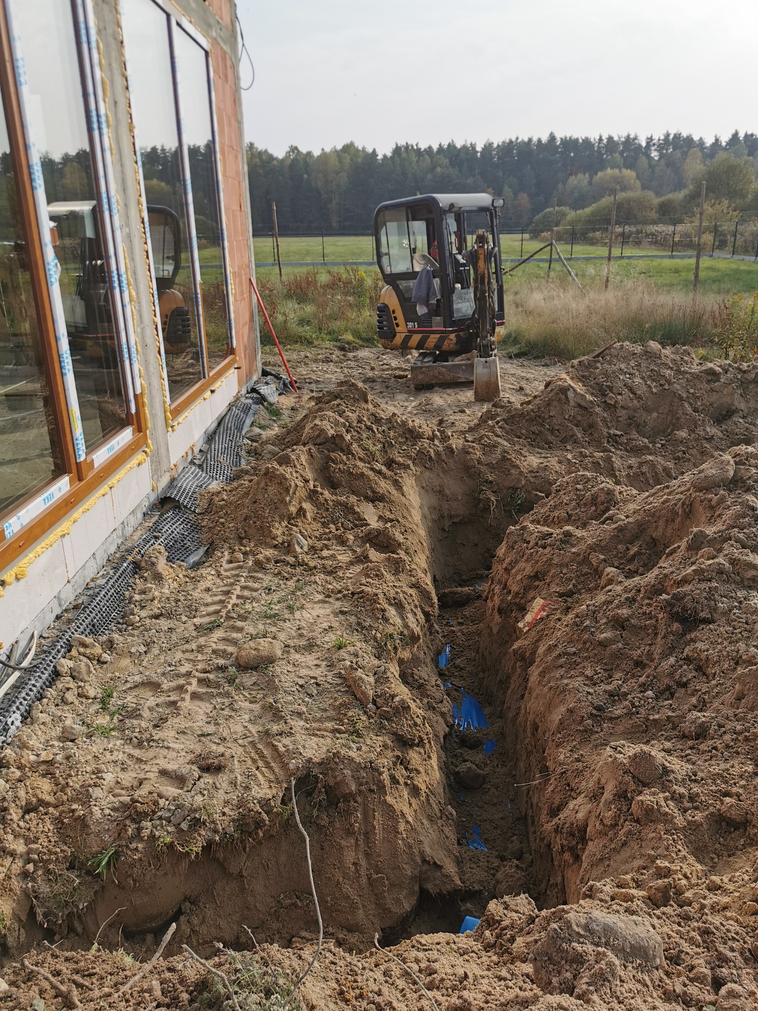 Wykopany rów przy fundamencie domu z widocznymi niebieskimi rurami wewnątrz, mała koparka stoi w tle, teren budowy.