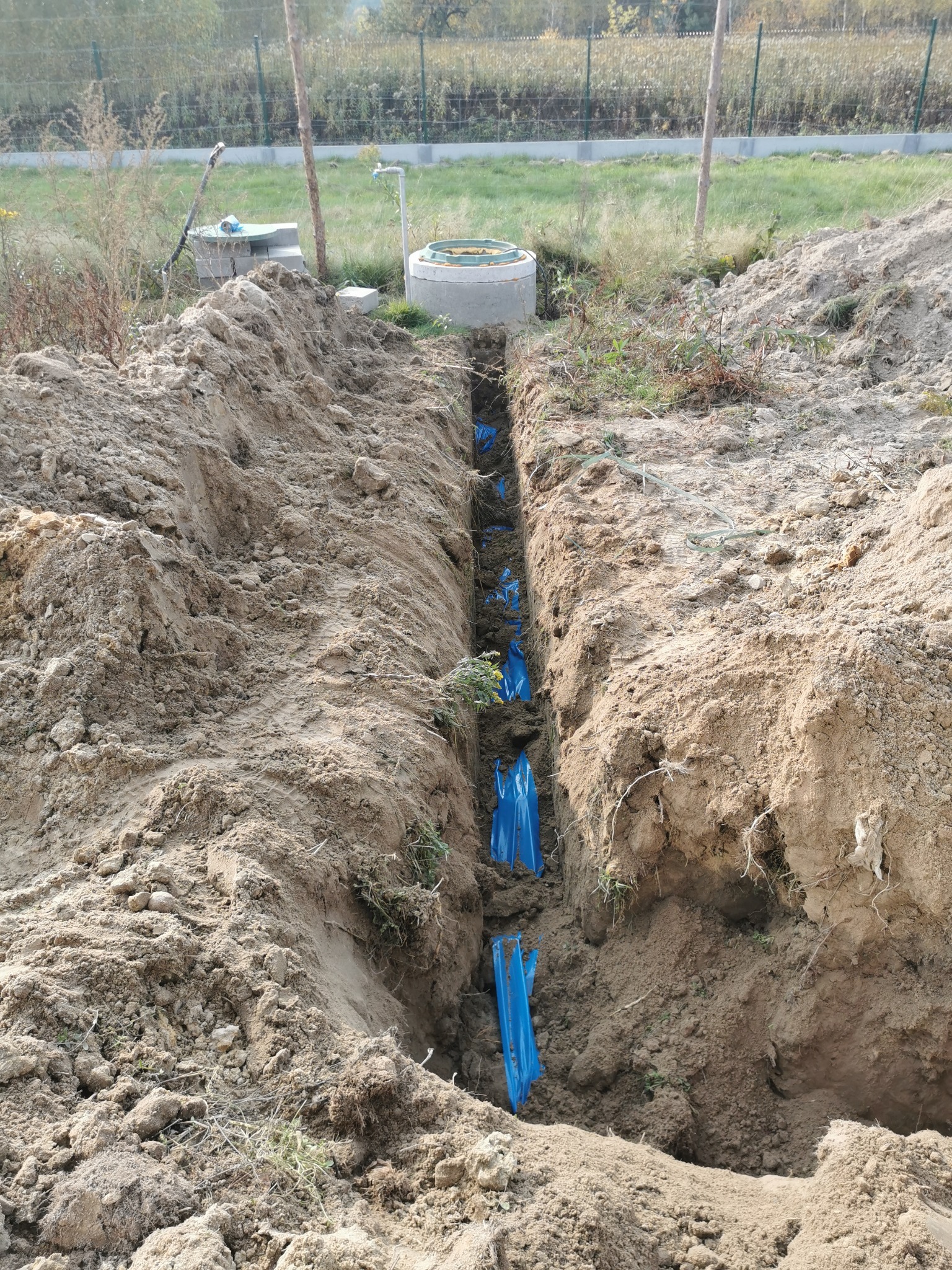 Wykopany wąski rów w ziemi z niebieskimi rurami w folii ochronnej, przygotowany pod instalację, widoczne elementy infrastruktury na działce.