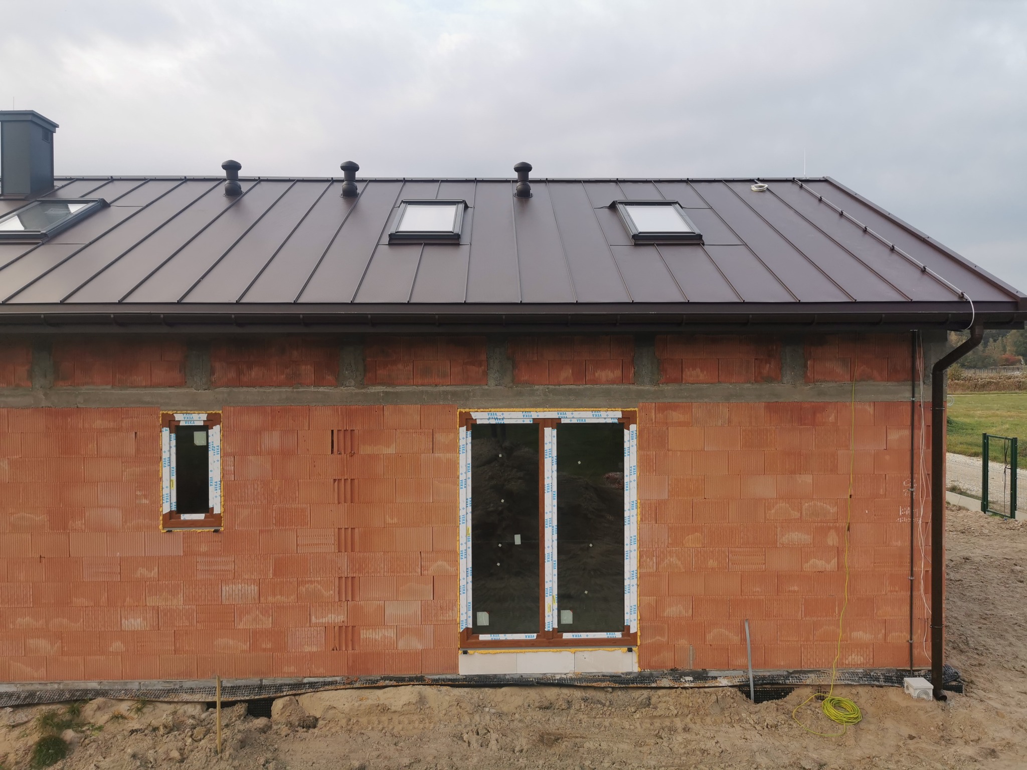 Niedokończony budynek z cegły ceramicznej z brązowym dachem, trzema oknami dachowymi, kominem i zamontowaną instalacją odgromową z żółtym przewodem uziemiającym.