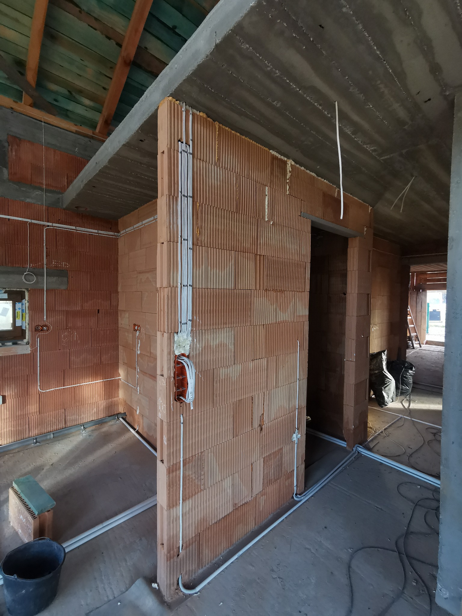 Wnętrze budynku w trakcie budowy z widoczną instalacją elektryczną w rurkach PCV, ściany z czerwonej cegły, surowy betonowy strop, widoczne kable i puszki instalacyjne.