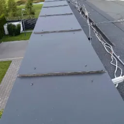 Szary dach z blachy na budynku jednorodzinnym, widok z góry na fragment połaci dachowej z widocznymi łączeniami blach i elementami mocującymi, w tle antena telewizyjna, drzewa i pola.