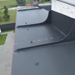 Szare, metalowe obróbki blacharskie na dachu z widocznymi śrubami mocującymi, na tle krajobrazu z drzewami i budynkami.