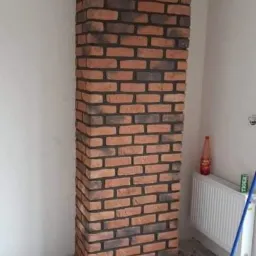 Wysoka kolumna z cegieł klinkierowych w trakcie montażu, widoczna na tle białej ściany, z wiadrem i narzędziami na podłodze.