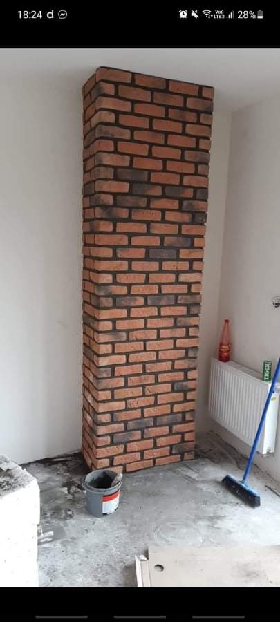 Wysoka kolumna z cegieł klinkierowych w trakcie montażu, widoczna na tle białej ściany, z wiadrem i narzędziami na podłodze.