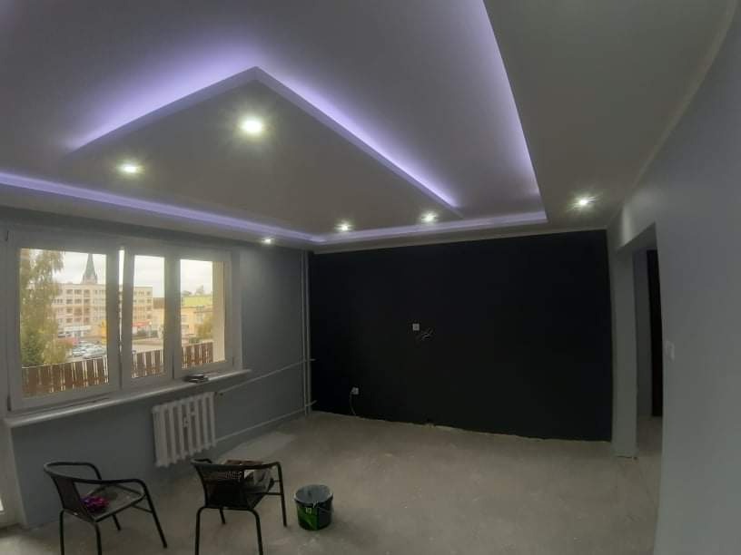 Pokój po remoncie z podwieszanym sufitem z oświetleniem LED, widok na miasto przez okno, dwa krzesła i wiadro po farbie na podłodze.