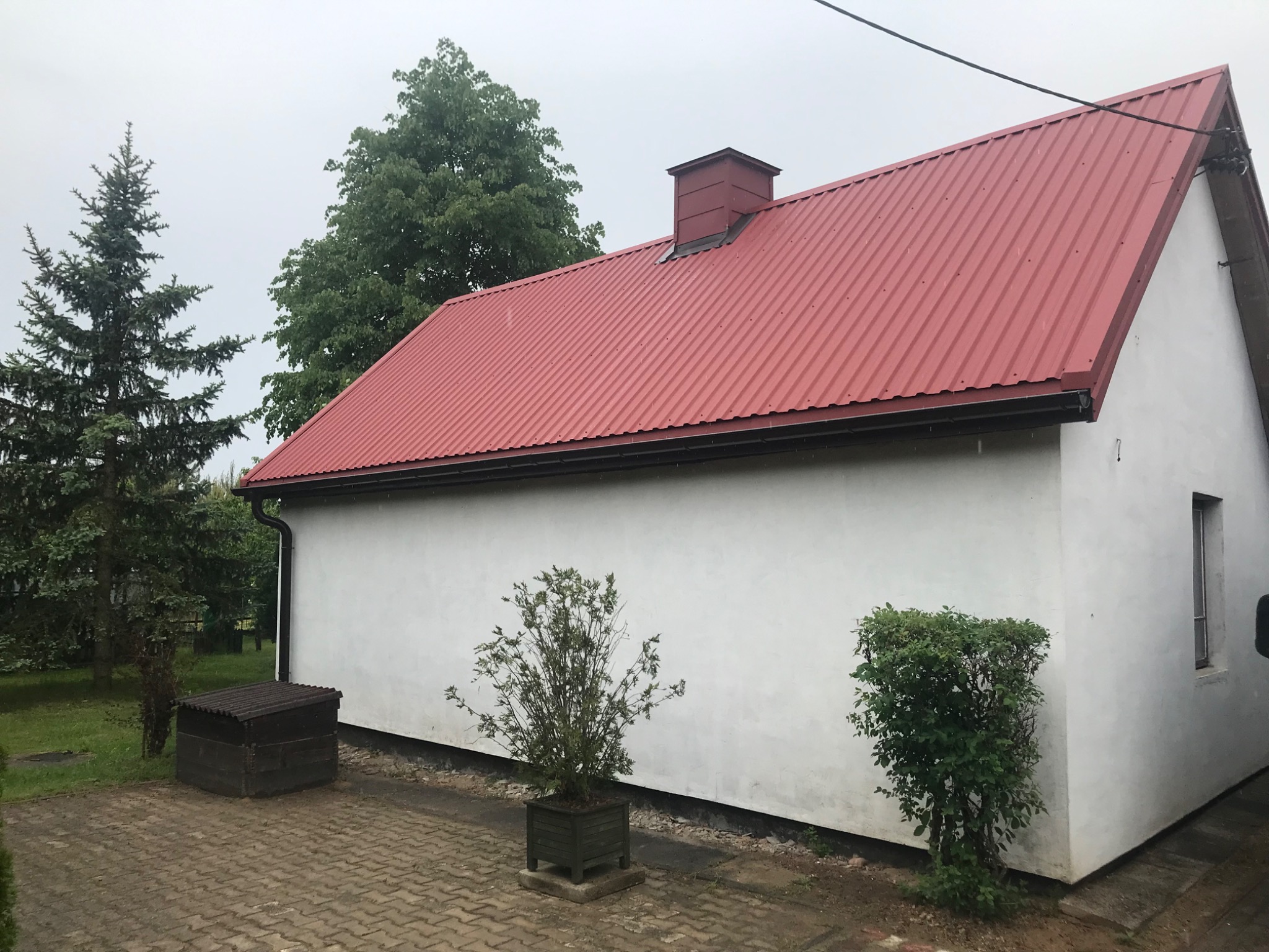 Dom z czerwonym, falistym dachem i kominem, widok z boku w pochmurny dzień. Widoczne drzewa i krzewy w otoczeniu.