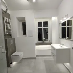 Nowoczesna, minimalistyczna łazienka w jasnych kolorach z podświetlaną toaletą, grzejnikiem drabinkowym i umywalką z szafką. W oknie widoczny biały storczyk.