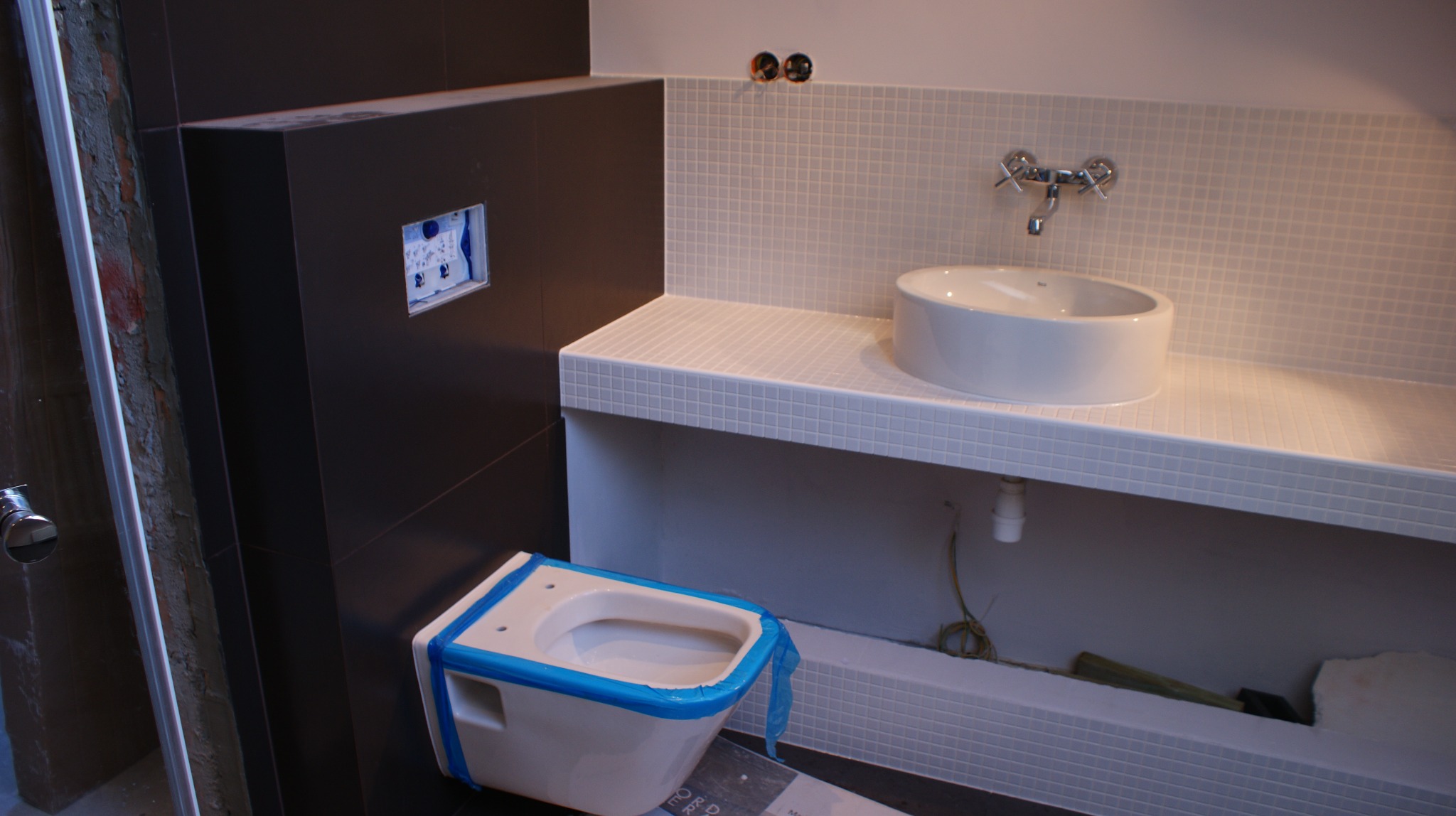 Niedokończona łazienka z umywalką nablatową na blacie z mozaiki, wisząca toaleta zabezpieczona niebieską taśmą malarską, widoczne instalacje i brakujące elementy wykończenia.