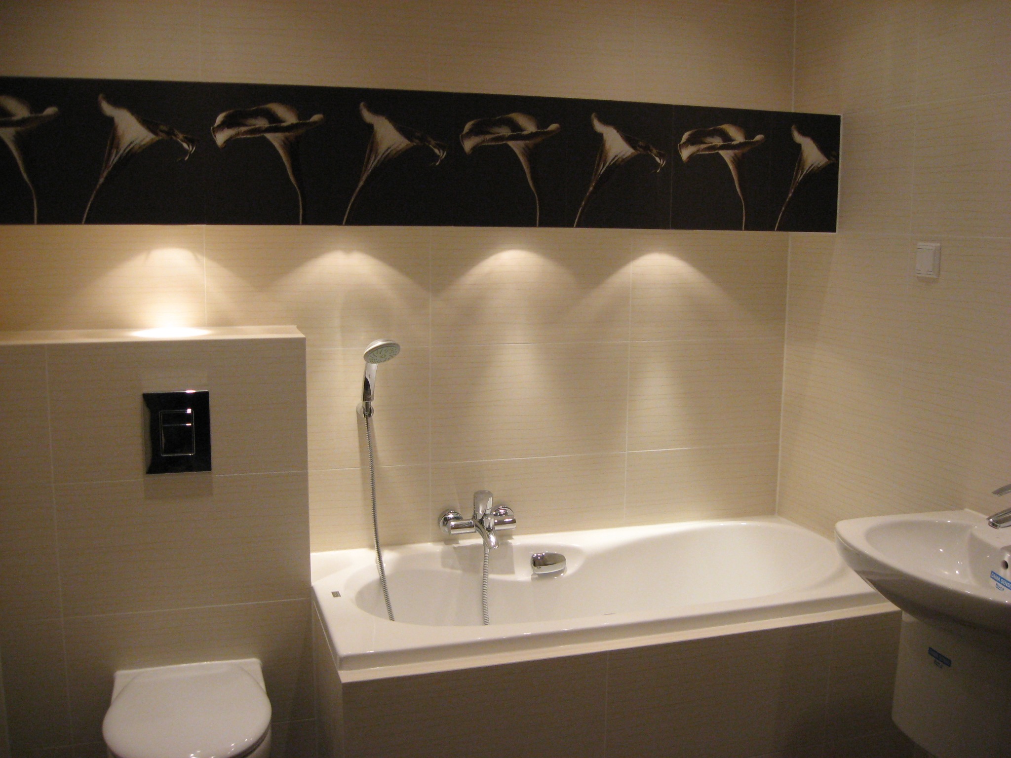 Wykończona łazienka z beżowymi płytkami, białą wanną i umywalką, ozdobnym panelem z motywem kwiatowym nad wanną oraz punktowym oświetleniem.