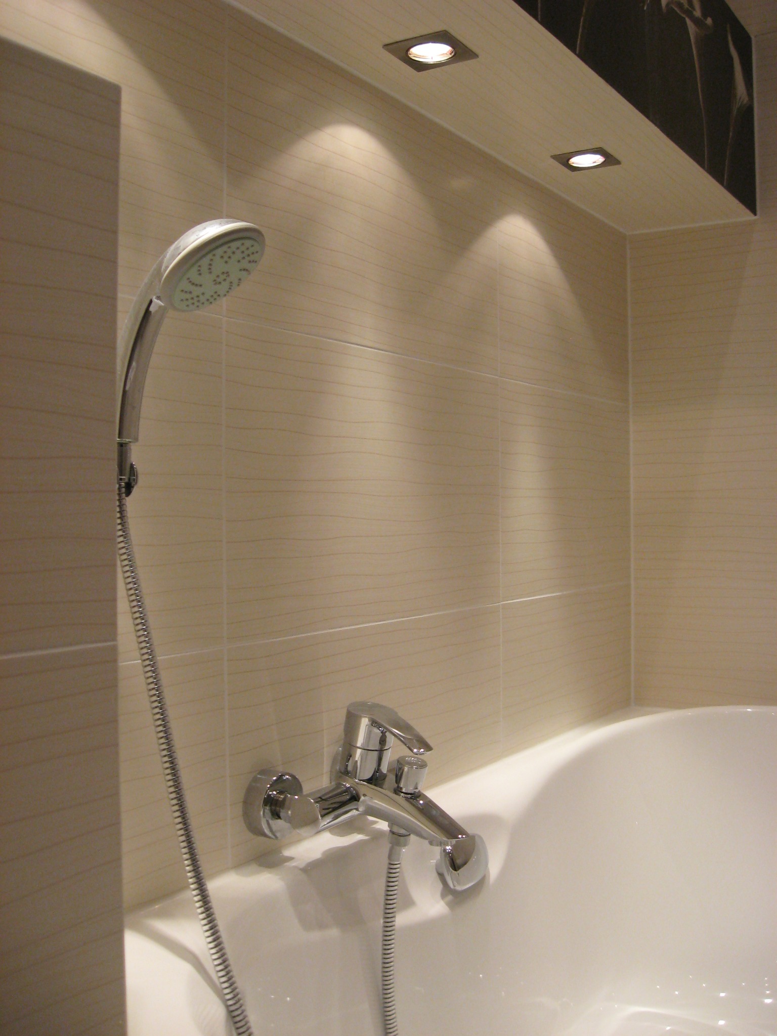 Biała wanna z chromowaną baterią i słuchawką prysznicową na tle beżowych płytek z delikatnym wzorem, oświetlona punktowymi lampami sufitowymi.