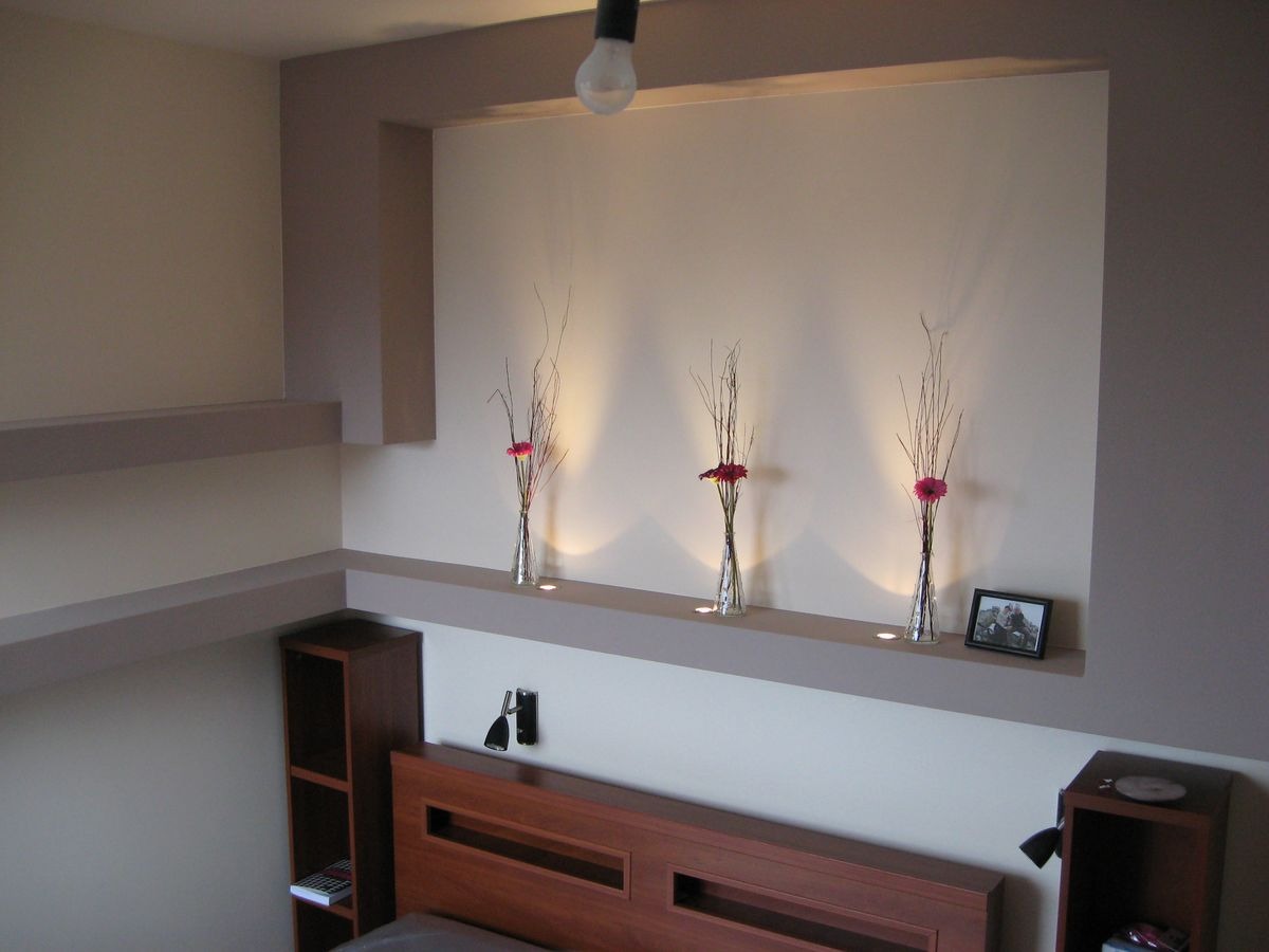 Wnęka w sypialni z trzema wazonami z kwiatami i oświetleniem punktowym, nad wezgłowiem łóżka z półkami i szafkami po bokach.