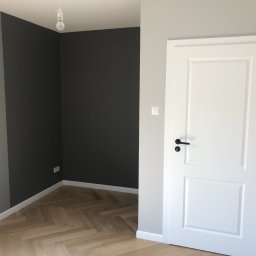 ReMax Usługi Remontowo-Budowlane - Wnętrze po remoncie: białe drzwi z czarną klamką, parkiet w jodełkę, ściany pomalowane na szaro i grafitowo, widoczna pusta żarówka