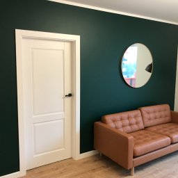 ReMax Usługi Remontowo-Budowlane - Nowoczesny salon z ciemnozielonymi ścianami, białymi drzwiami z czarną klamką, okrągłym lustrem w złotej ramie i brązową skórzaną sofą na drewnianej podłodze.