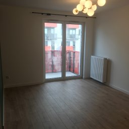 ReMax Usługi Remontowo-Budowlane - Puste, jasne pomieszczenie z panelami podłogowymi, grzejnikiem i nowoczesną lampą sufitową w stylu atomu, widok na balkon z pomarańczową siatką zabezpieczającą.