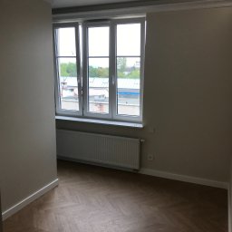 ReMax Usługi Remontowo-Budowlane - Pusty pokój po remoncie z parkietem w jodełkę, białymi ścianami z listwami przypodłogowymi i oknem z widokiem na budynki.