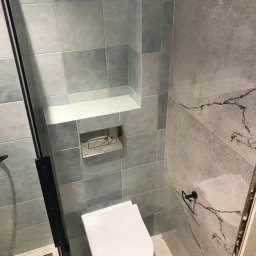 ReMax Usługi Remontowo-Budowlane - Nowoczesna łazienka po remoncie z płytkami w odcieniach szarości i błękitu, wisząca toaleta z chromowanym przyciskiem spłukiwania, wnęka w ścianie wyłożona białymi płytkami, czarny wieszak na ręcznik.