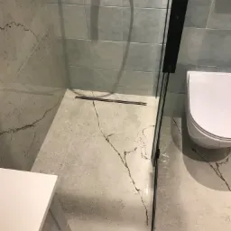 Nowoczesna łazienka z szarymi płytkami imitującymi marmur, kabiną prysznicową typu walk-in z czarnymi profilami i odpływem liniowym, oraz podwieszaną toaletą.