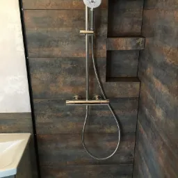 Kabina prysznicowa z deszczownicą i półką w ścianie wyłożonej płytkami imitującymi rdzewiejący metal, widoczny fragment umywalki.