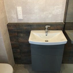 ReMax Usługi Remontowo-Budowlane - Umywalka nablatowa z szarą szafką, chromowaną baterią i odpływem, zamontowana na tle ściany z płytek imitujących drewno, obok toaleta i lustro.