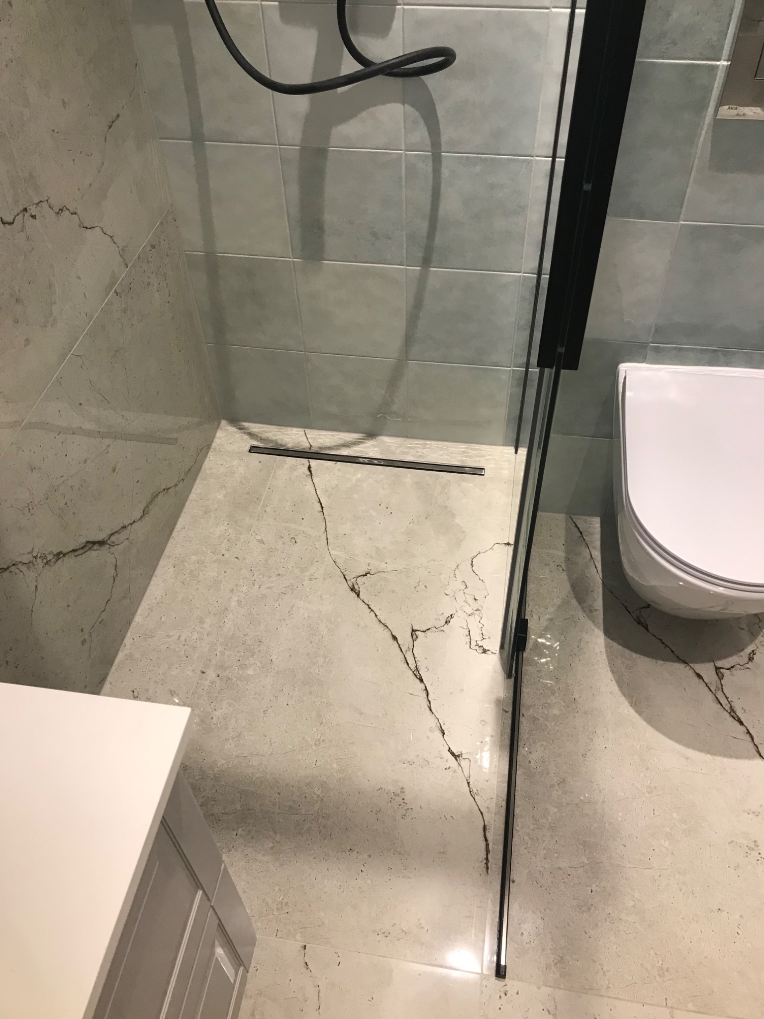 Nowoczesna łazienka z szarymi płytkami imitującymi marmur, kabiną prysznicową typu walk-in z czarnymi profilami i odpływem liniowym, oraz podwieszaną toaletą.
