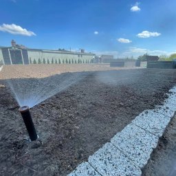 Zielony Ogródek Rafał Szmelich - Automatyczny system nawadniania w trakcie pracy na przygotowanym terenie pod trawnik, widoczne zraszacze, obrzeża z kostki, budynki w tle i błękitne niebo.