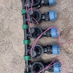 Zielony Ogródek Rafał Szmelich - Bliski widok na czarny kolektor z zielonymi złączkami i niebieskimi końcówkami, z podłączonymi przewodami elektrycznymi, leżący na żwirowej powierzchni.