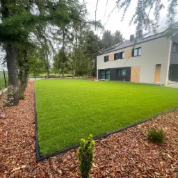 Nowoczesny ogród z intensywnie zielonym trawnikiem, obrzeżem z tworzywa i korą drzewną, na tle domu z elementami drewnianymi i zielenią drzew.