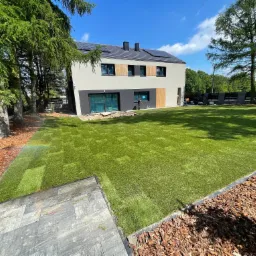 Trawa z rolki ułożona na posesji z nowoczesnym domem. System nawadniania w trakcie pracy, tworzący tęczę. Kora drzewna jako element dekoracyjny rabat.