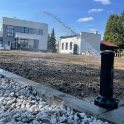 Automatyczny system nawadniania w akcji: zraszacz rozpyla wodę na przygotowany grunt, obok widoczny kamienny obrzeże trawnika i budynki w tle, błękitne niebo.