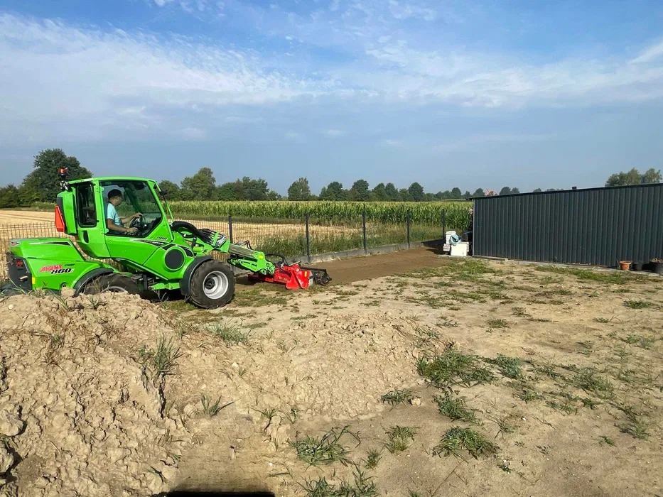 Operator w zielonej maszynie Avant 860 wyrównuje teren pod przyszły trawnik, widoczny świeżo przygotowany pas ziemi wzdłuż ogrodzenia.