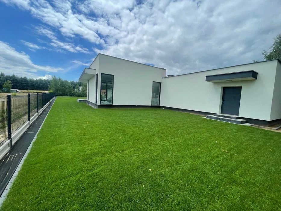 Nowoczesny dom z białymi ścianami i idealnie przystrzyżonym trawnikiem, ogrodzony czarnym metalowym płotem na tle błękitnego nieba z chmurami.