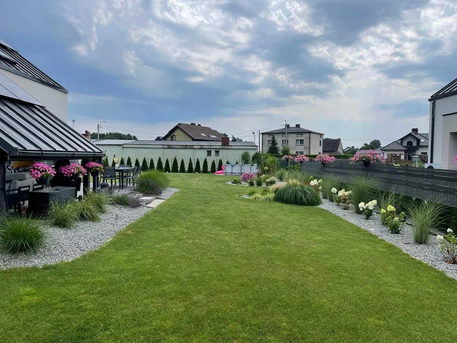 Rozległy, zadbany trawnik z dekoracyjnymi nasadzeniami wzdłuż ogrodzenia i ścieżek wysypanych żwirem, w tle budynki mieszkalne i basen z dmuchanym flamingiem.