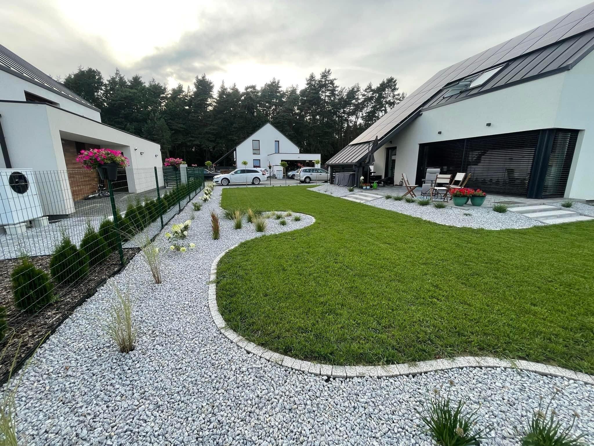 Nowoczesny ogród z równo przystrzyżonym trawnikiem, ścieżką z białego żwiru obramowaną kostką, ozdobnymi krzewami i kwiatami oraz nowoczesnymi domami w tle.