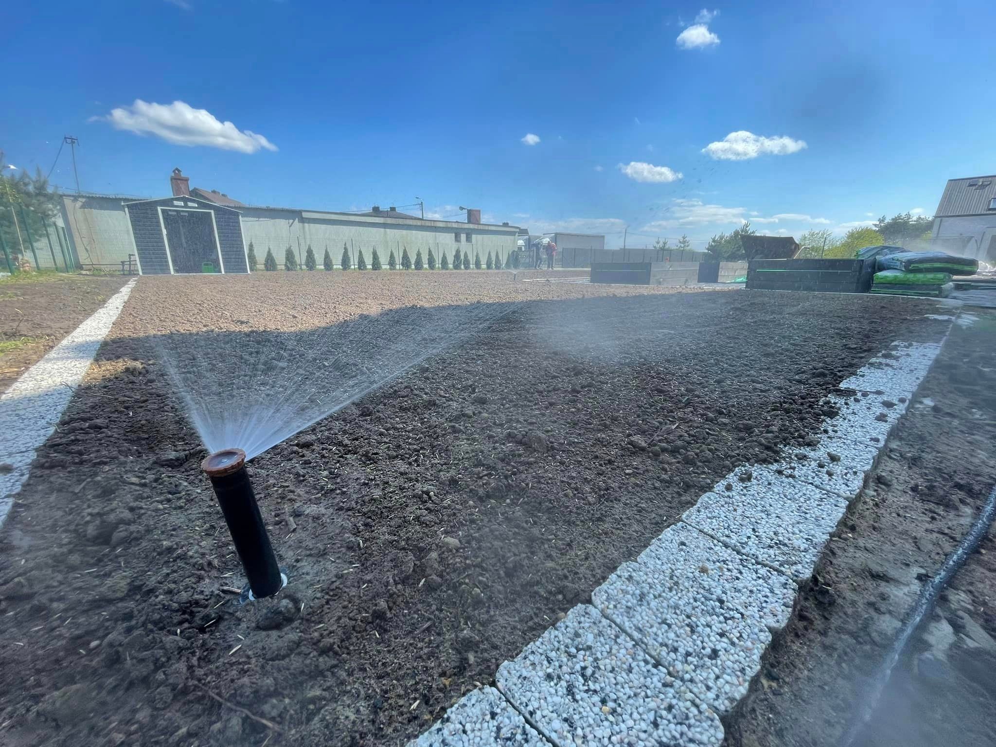 Automatyczny system nawadniania w trakcie pracy na przygotowanym terenie pod trawnik, widoczne zraszacze, obrzeża z kostki, budynki w tle i błękitne niebo.